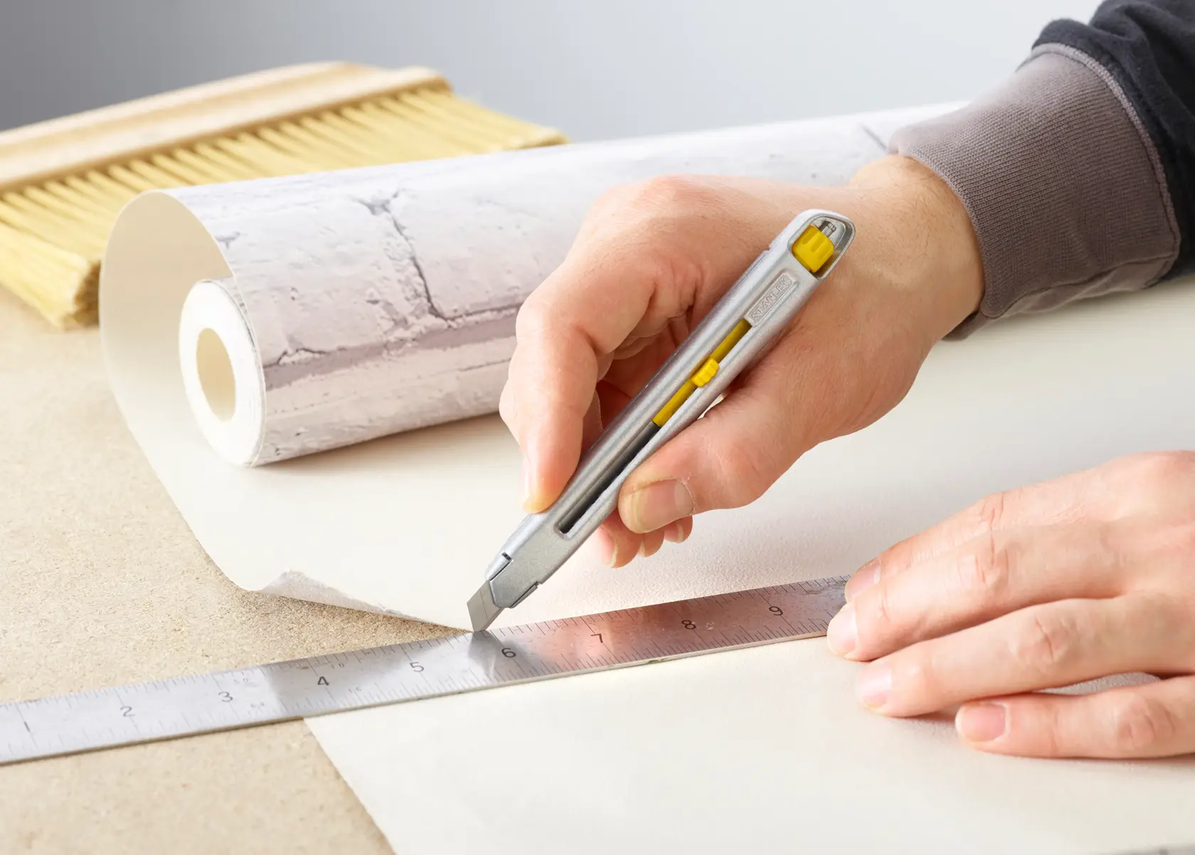 Photo of STANLEY 0-10-095 utility knife cutting wallpaper along a metal ruler, with wallpaper roll and brush visible.