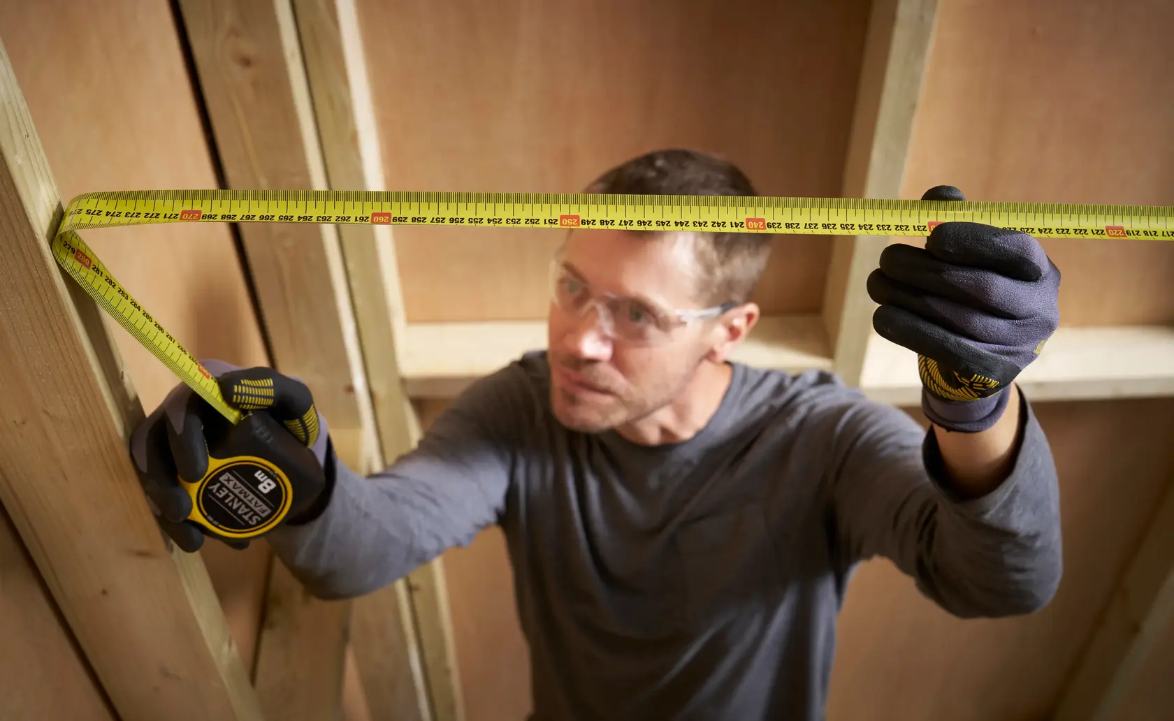 Photo of a man measuring wood framing with a STANLEY 0-33-728 tape measure, wearing gloves and safety glasses.