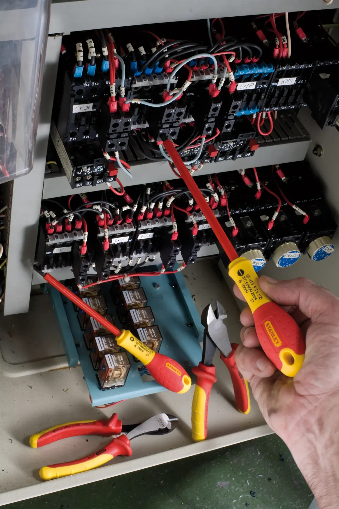 STANLEY 0-65-413 insulated screwdriver in hand near electrical panel, pliers and tools visible, angled from above.