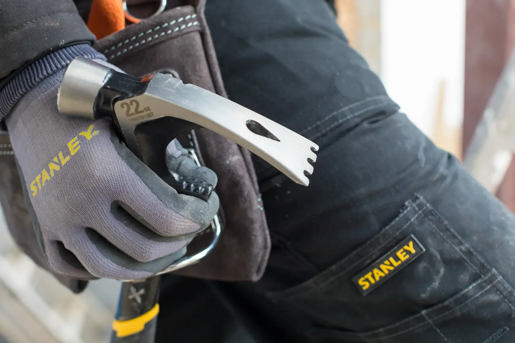 STANLEY FMHT51367-2 hammer held by a gloved hand beside work trousers with STANLEY logo, close-up view.