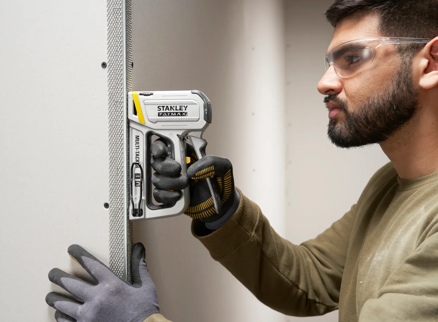 Photo of STANLEY FATMAX FMHT70250-0 stapler in use on drywall, held by gloved hands during installation.
