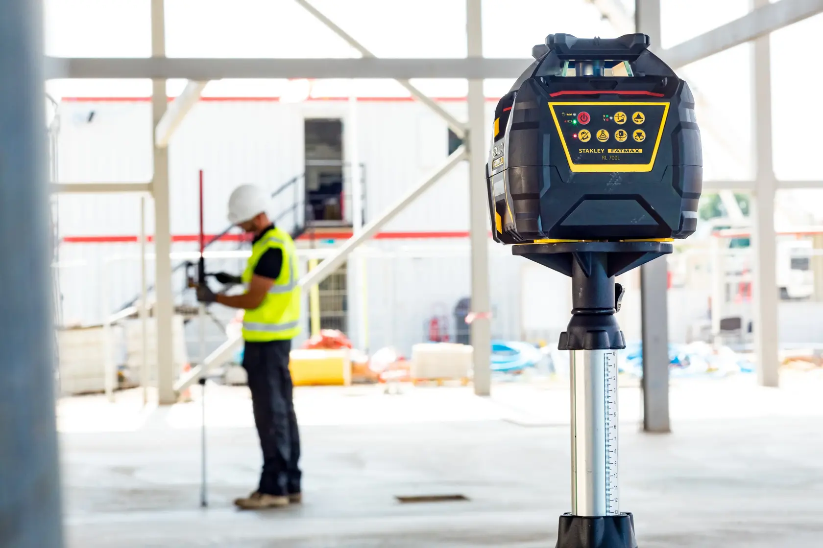 STANLEY FMHT77449-1 rotary laser level on tripod at construction site, worker in background.
