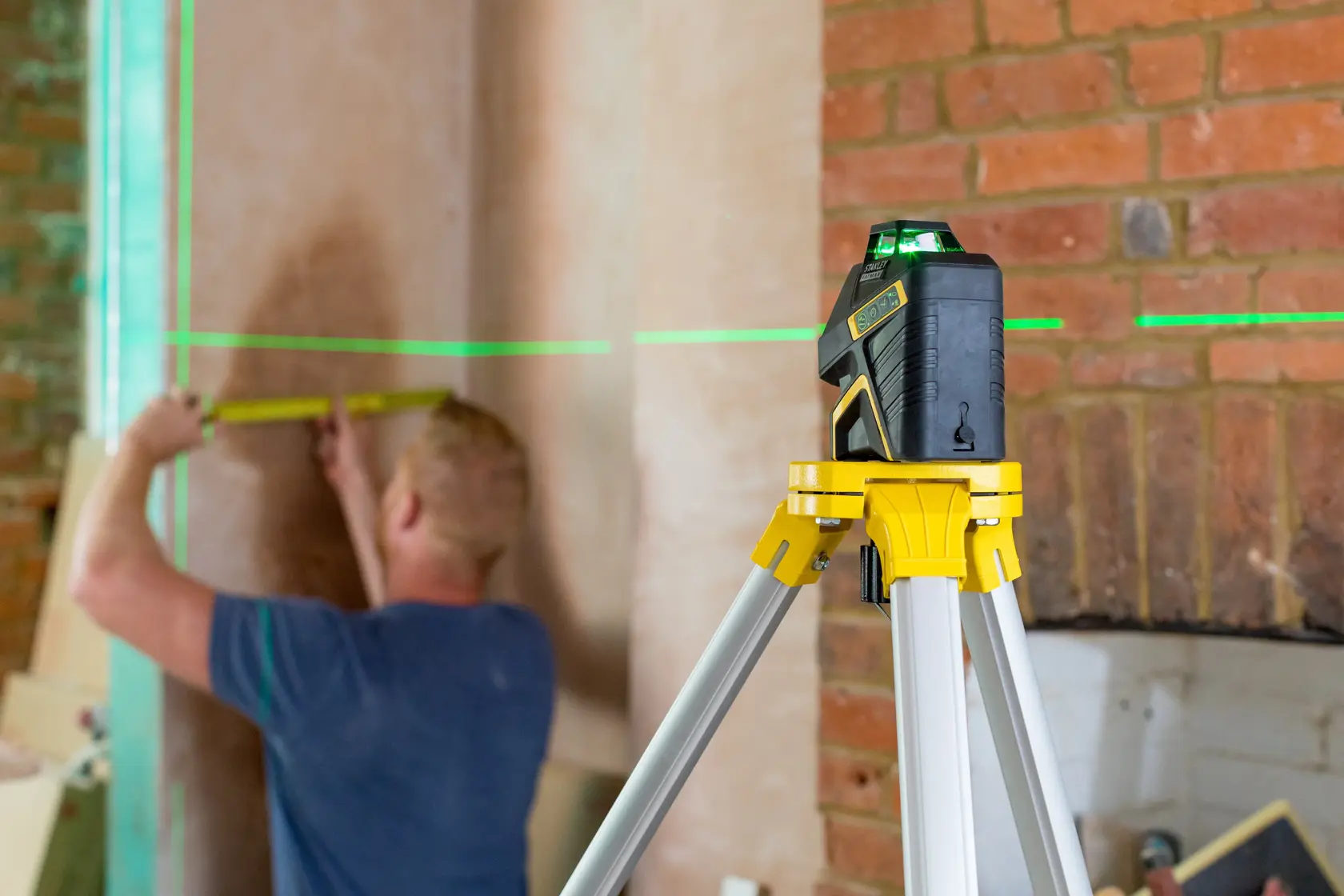 STANLEY FMHT77617-1 laser level on tripod projects green lines while person measures wall in room.