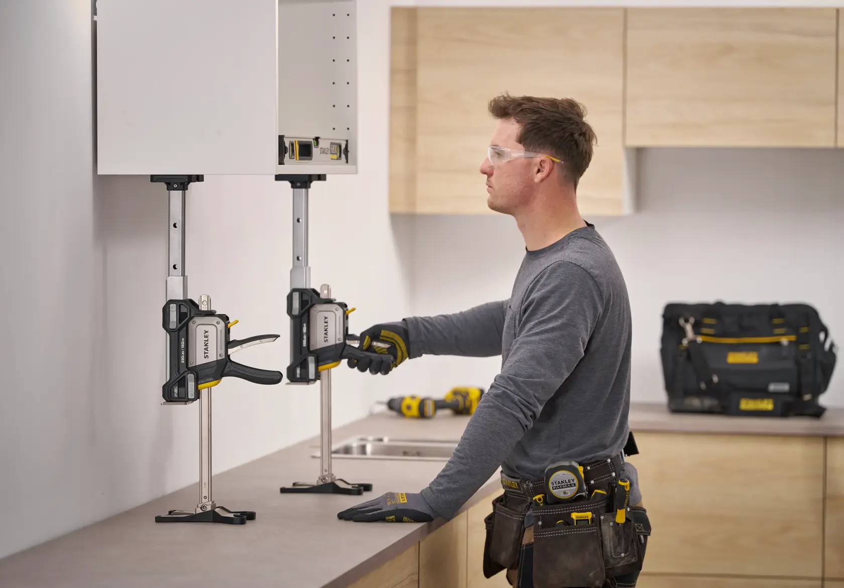 STANLEY FMHT83554-1 cabinet clamp holding wall cabinet during installation, with worker adjusting clamp in kitchen setting.
