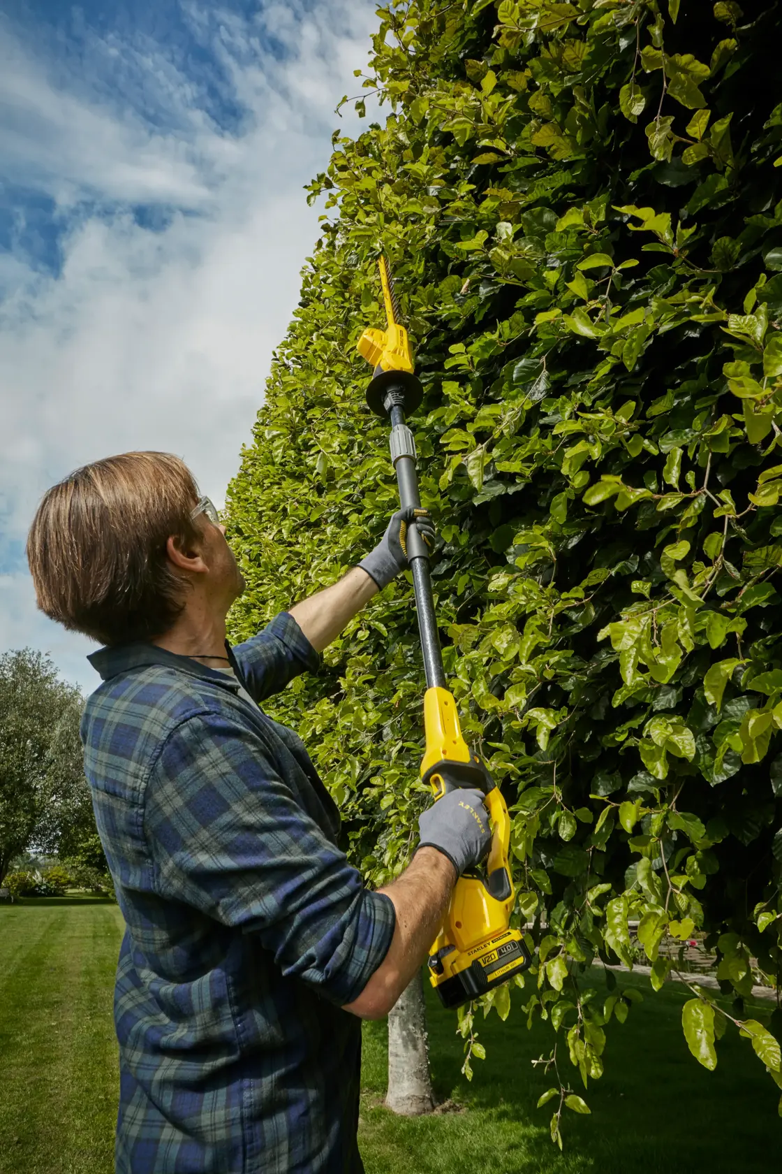 STANLEY SFMCPH845M1 pole hedge trimmer in use, person trimming tall green hedge outdoors.