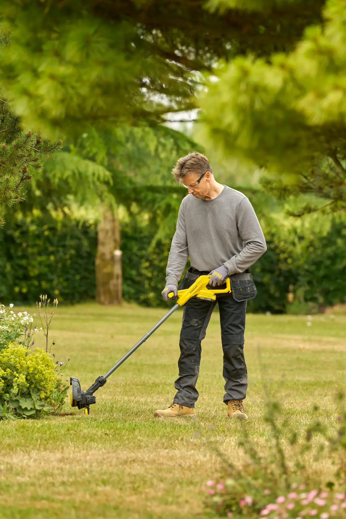 STANLEY SFMCST925B string trimmer used by a person trimming grass in a garden, side outdoor view.