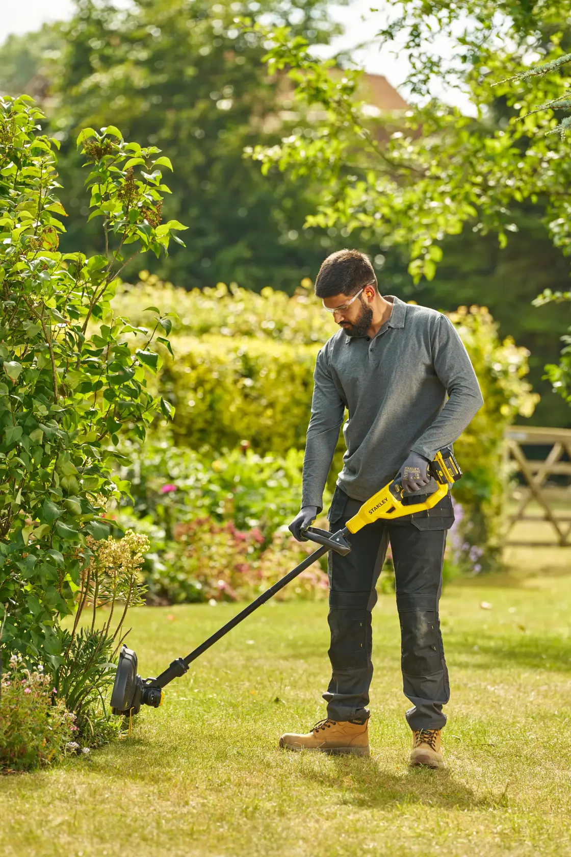 Photo of a person using a STANLEY STCST933B grass trimmer in a garden, holding the yellow and black tool on green grass.