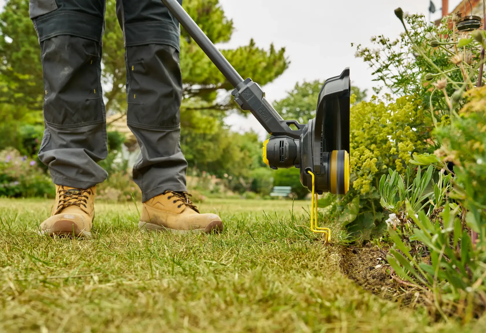 STANLEY STCST933M1 grass trimmer in use, side view, trimming lawn edge near garden plants.