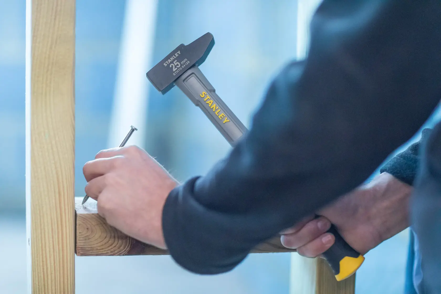 Photo of STANLEY STHT0-54129 hammer driving nail into wooden beam, close-up of worker’s hands.
