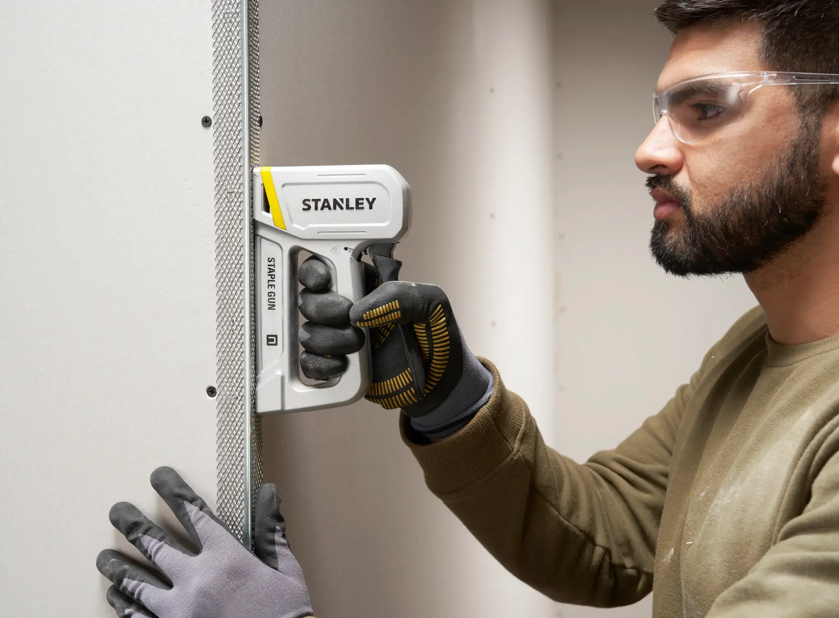 Photo of STANLEY STHT70150-0 staple gun in use, held against drywall by a gloved hand.